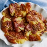 A white plate loaded with crispy smashed potatoes, sprinkled with golden seasoning, is set on a white surface. Theres a light blue cloth hanging out in the background.