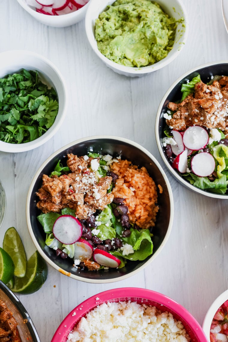 A bowl of al pastor salad—think Mexican rice, juicy meat, lettuce, black beans, radishes, and cheese—sits on a white table. Around it are bowls with guac, fresh cilantro, lime wedges, and extra cheese for topping.