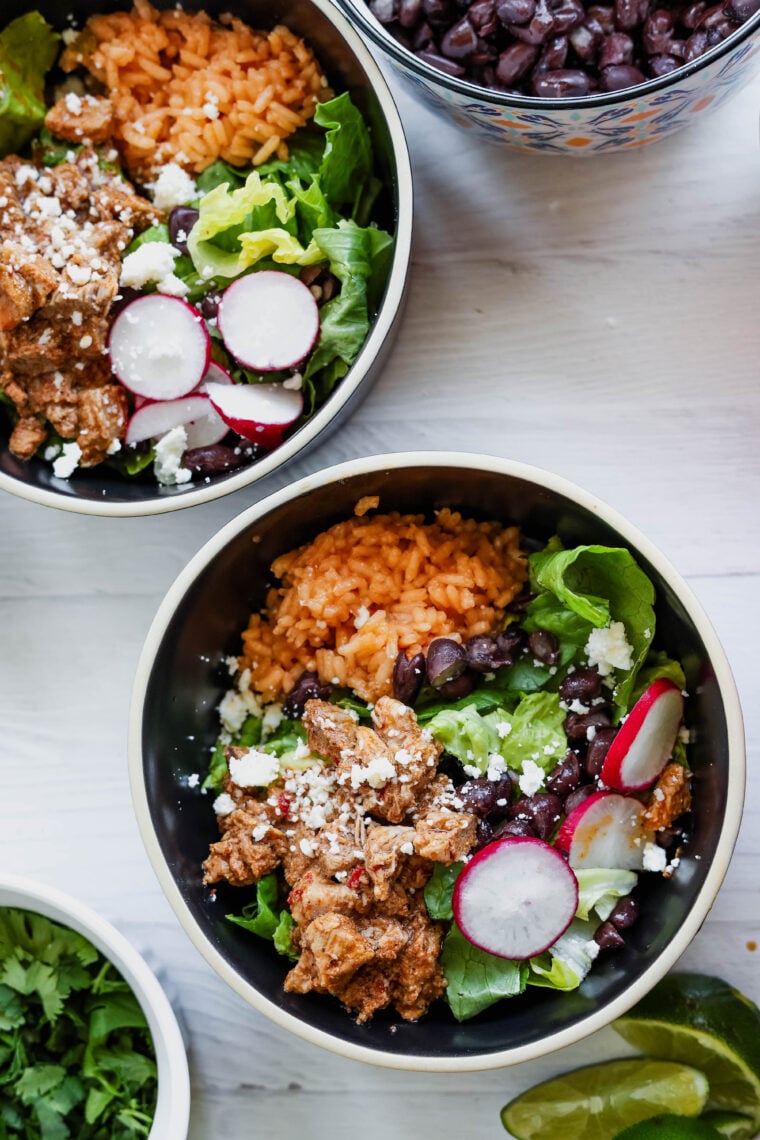 Two bowls packed with tasty al pastor salad, orange rice, black beans, lettuce, radishes, and crumbled cheese. There’s also a bowl of black beans and some lime wedges on the side—all sitting on a white countertop.
