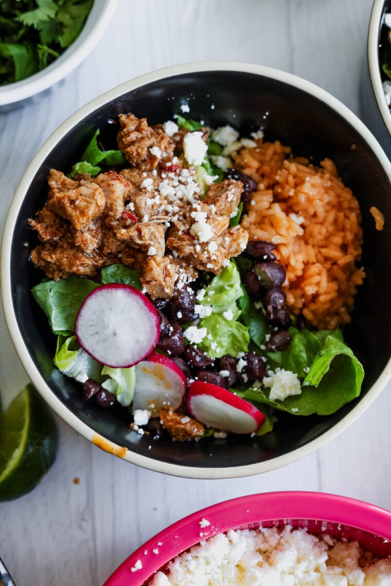 A bowl packed with spiced ground meat, orange rice, black beans, lettuce, radishes, and crumbled cheese—this al pastor salad comes with a lime wedge on the side. You can see more tasty bowls in the background too.