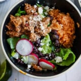 A bowl packed with spiced ground meat, orange rice, black beans, lettuce, radishes, and crumbled cheese—this al pastor salad comes with a lime wedge on the side. You can see more tasty bowls in the background too.