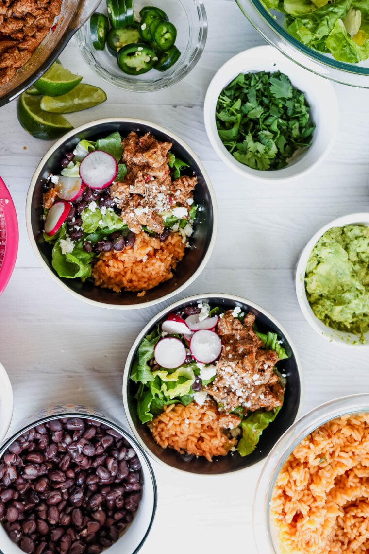 Two bowls of al pastor salad—packed with Mexican rice, lettuce, black beans, seasoned meat, radishes, and cheese—are chilling on a white table. Around them: guac, cilantro, jalapeños, lime wedges, extra black beans, and more rice.
