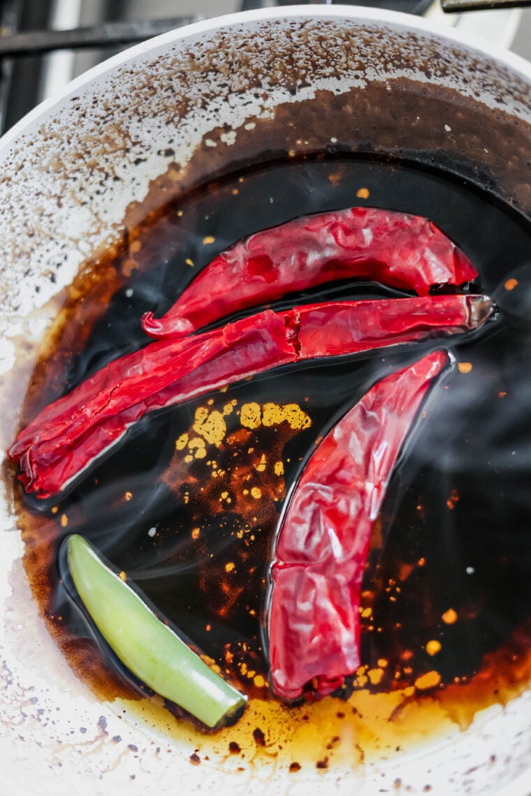 Three dried red chilis and a piece of green chili are simmering in a dark sauce in a white pan, with oil and sauce splatters on the sides. They’re just what you need to give your al pastor salad some serious kick!.