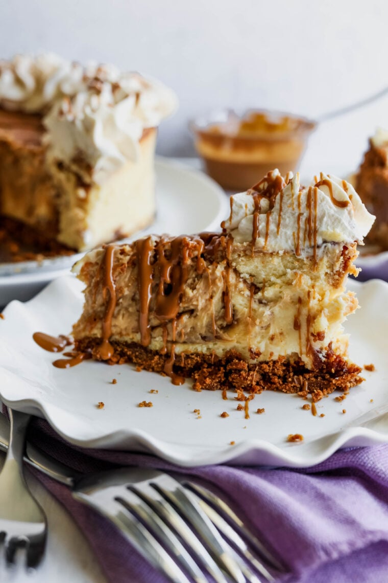 A slice of creamy biscoff white chocolate cheesecake sits on a white plate, topped with whipped cream and caramel drizzle, with some crumbs and silver forks nearby. You can see the rest of the cheesecake looking all blurry in the background.