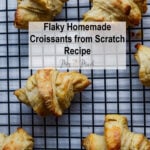 Freshly baked, golden-brown croissants cooling on a wire rack. A clear box says, “Flaky Homemade Croissants from Scratch Recipe” with “Mrs. Madi” in script. Perfect for anyone who loves croissants!.