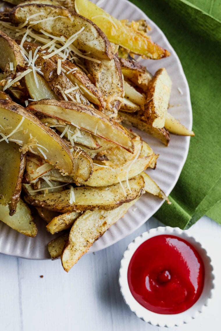 A plate of cheesy baked potato wedges is hanging out next to a little bowl of ketchup on a white table, and there’s a green napkin close by.