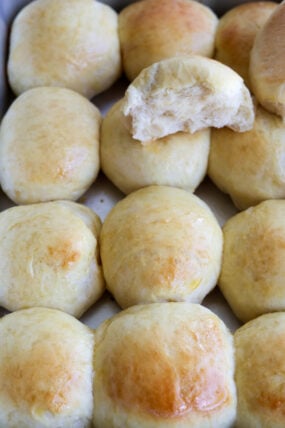 A close-up shot of a bunch of shiny, golden-brown dinner rolls—one at the top has a bite taken out, showing off its super soft, fluffy inside.
