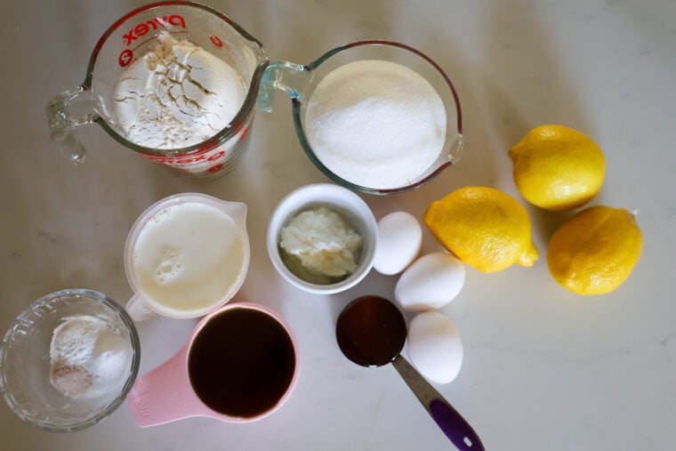 Looking down at a white countertop with all the baking essentials—flour, sugar, three lemons, eggs, milk, some brown liquid (honey or syrup?), shortening, baking powder, and a cup of white powder. Ready to whip something up!.