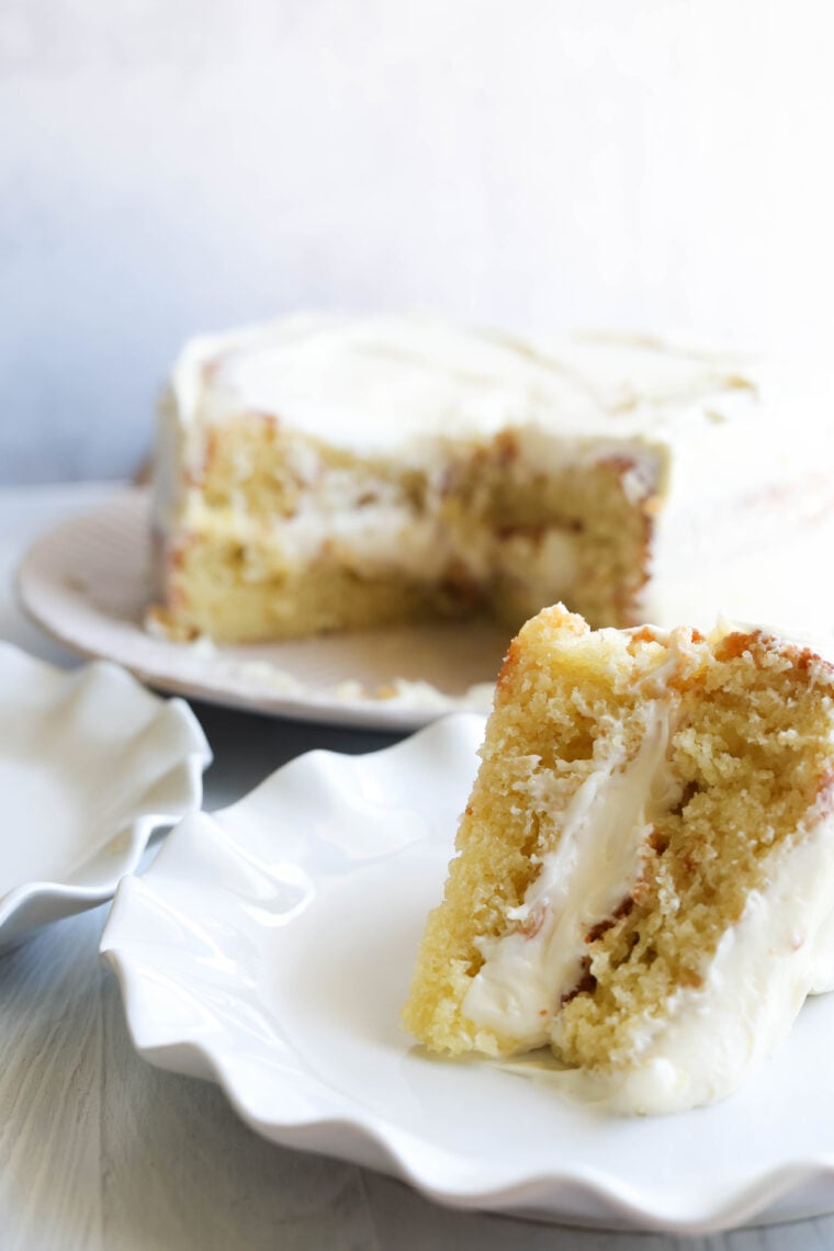 Theres a slice of yellow cake with white frosting and creamy filling on a white plate, and you can see the rest of the cake chilling in the background on a cake stand.