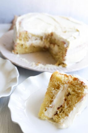 Theres a slice of yellow cake with white frosting on a plate up front, and you can see the rest of the cake—missing a slice—sitting in the background.