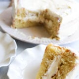 Theres a slice of yellow cake with white frosting on a plate up front, and you can see the rest of the cake—missing a slice—sitting in the background.