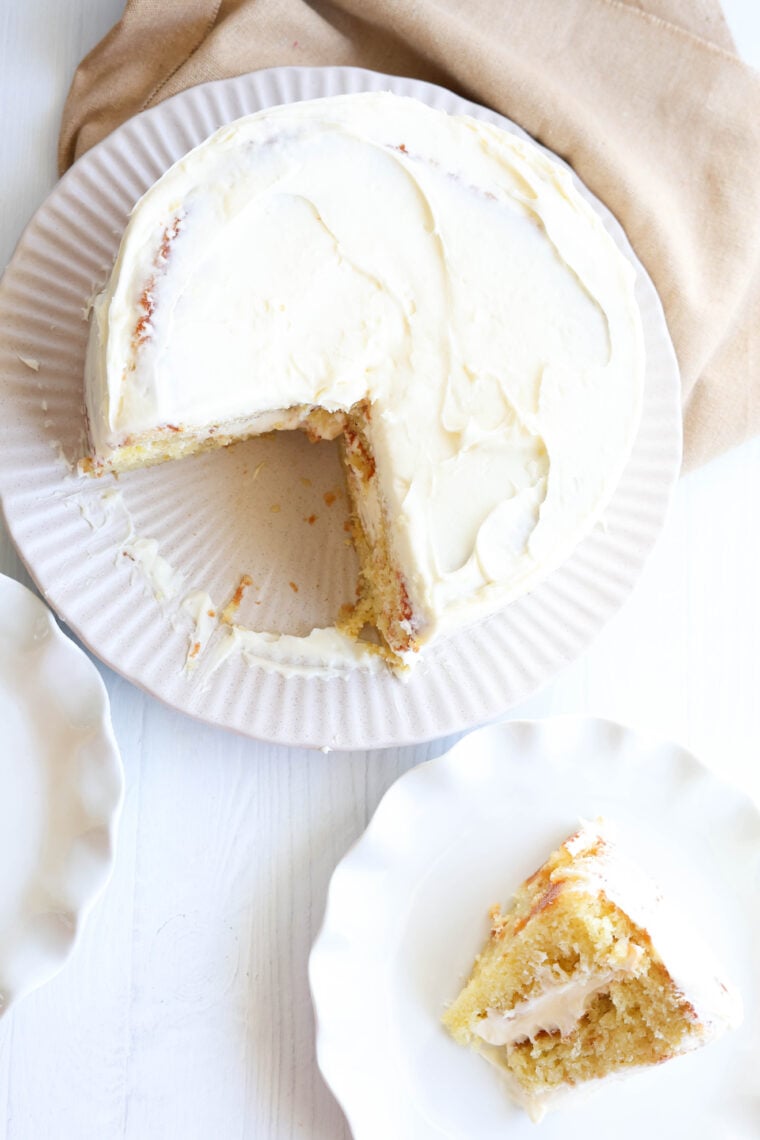 A round, frosted cake with a slice missing is on a white plate. The missing piece is hanging out on a smaller plate next to it. Everything’s sitting on a white surface with a beige cloth in the background.