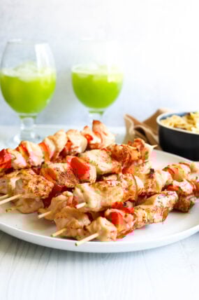 Skewers loaded with chicken and red bell peppers sit on a white plate, with a bowl of pasta and two glasses of green drinks in the background.