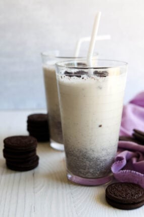 Two tall glasses of cookies and cream milkshake with white straws, topped with crushed cookies. Stacks of chocolate sandwich cookies are next to the glasses on a light surface, with a purple cloth nearby.