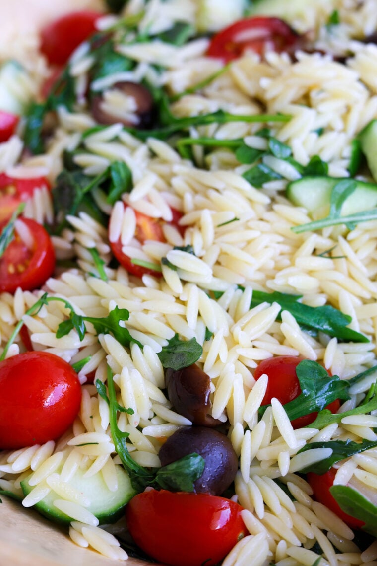 Heres a close-up of a super fresh orzo pasta salad packed with cherry tomatoes, cucumbers, black olives, arugula, and parsley. All the bright colors and flavors mixed together make this dish look extra tasty!.