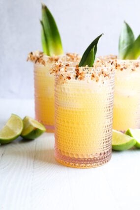A cool glass packed with a bright yellow tropical drink, topped off with toasted coconut on the rim and a pineapple leaf. There are some lime wedges next to it, plus two more drinks just like it in the background.