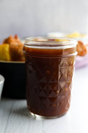 A glass jar of dark brown BBQ sauce sits on a white surface, with some food and bowls blurry in the background.