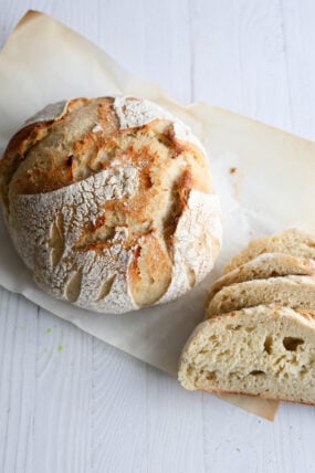 A round, rustic loaf of bread with a golden, crispy crust sits on parchment paper, dusted with flour. Next to it are a few thick slices, all set on a white wooden table.
