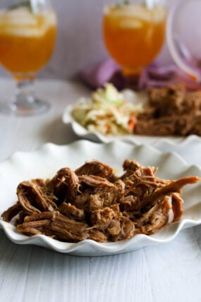 Pulled pork piled on a white plate, coleslaw chilling in the back, and two iced drinks ready to go.