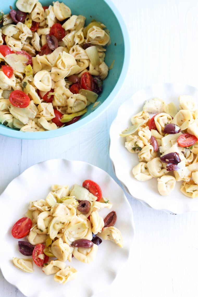 A blue bowl and two white plates filled with tortellini pasta salad—loaded with cherry tomatoes, artichoke hearts, olives, and herbs—sit on a white surface.
