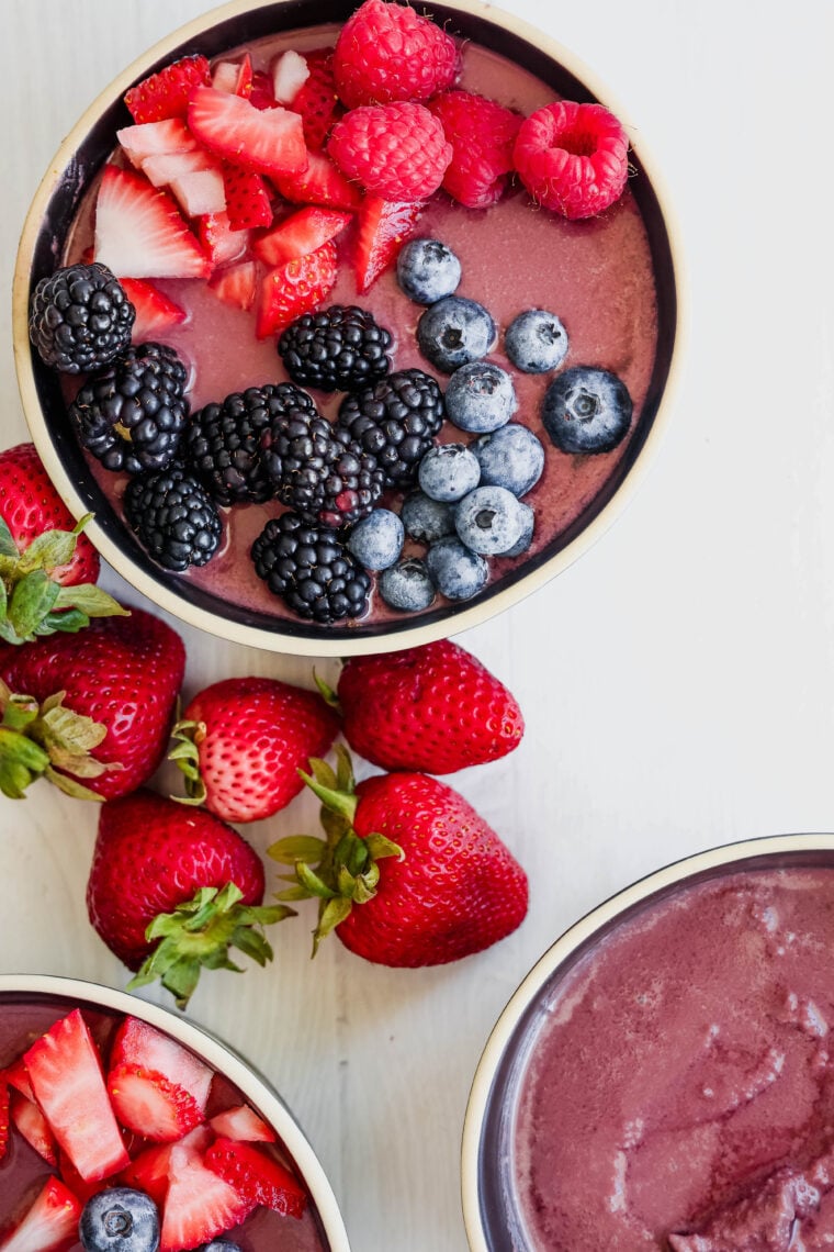 A tasty acai smoothie bowl topped with sliced strawberries, raspberries, blackberries, and blueberries. Fresh strawberries and other bowls are scattered around on a white surface.