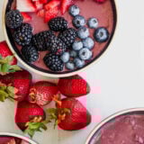 A tasty acai smoothie bowl topped with sliced strawberries, raspberries, blackberries, and blueberries. Fresh strawberries and other bowls are scattered around on a white surface.