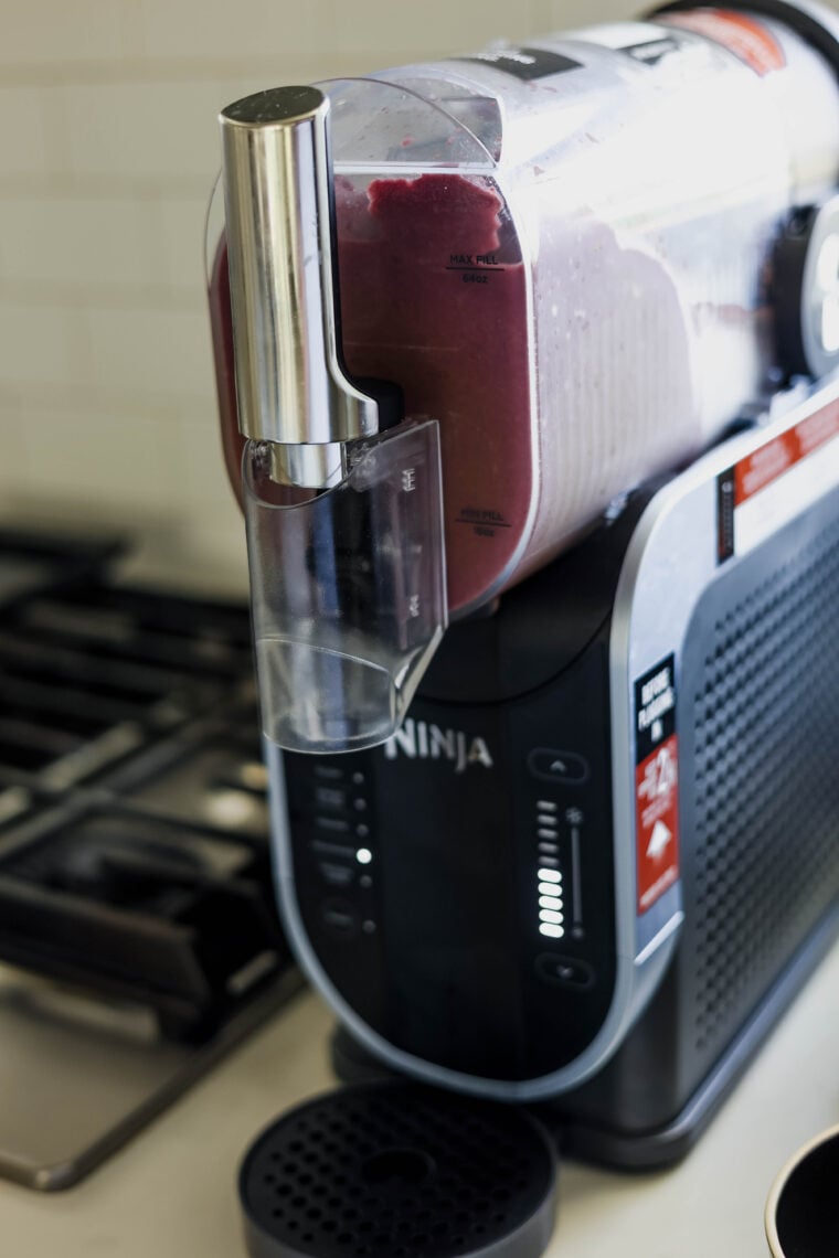 A Ninja smoothie maker sits on the counter, pouring a thick, purple acai bowl smoothie into a cup. Its digital screen is glowing, and you can spot the stove in the background.