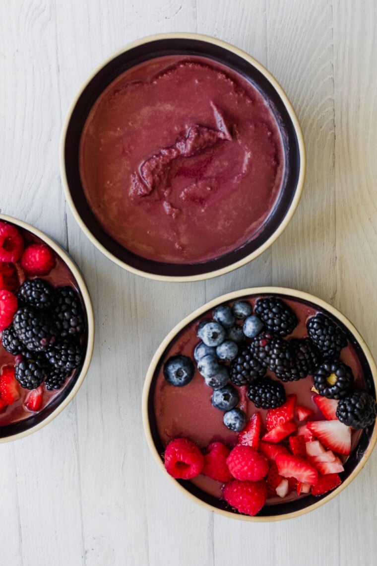 Three bowls sit on a light wooden table—one’s a smooth purple acai bowl and the other two are loaded with fresh berries like raspberries, blackberries, blueberries, and sliced strawberries.