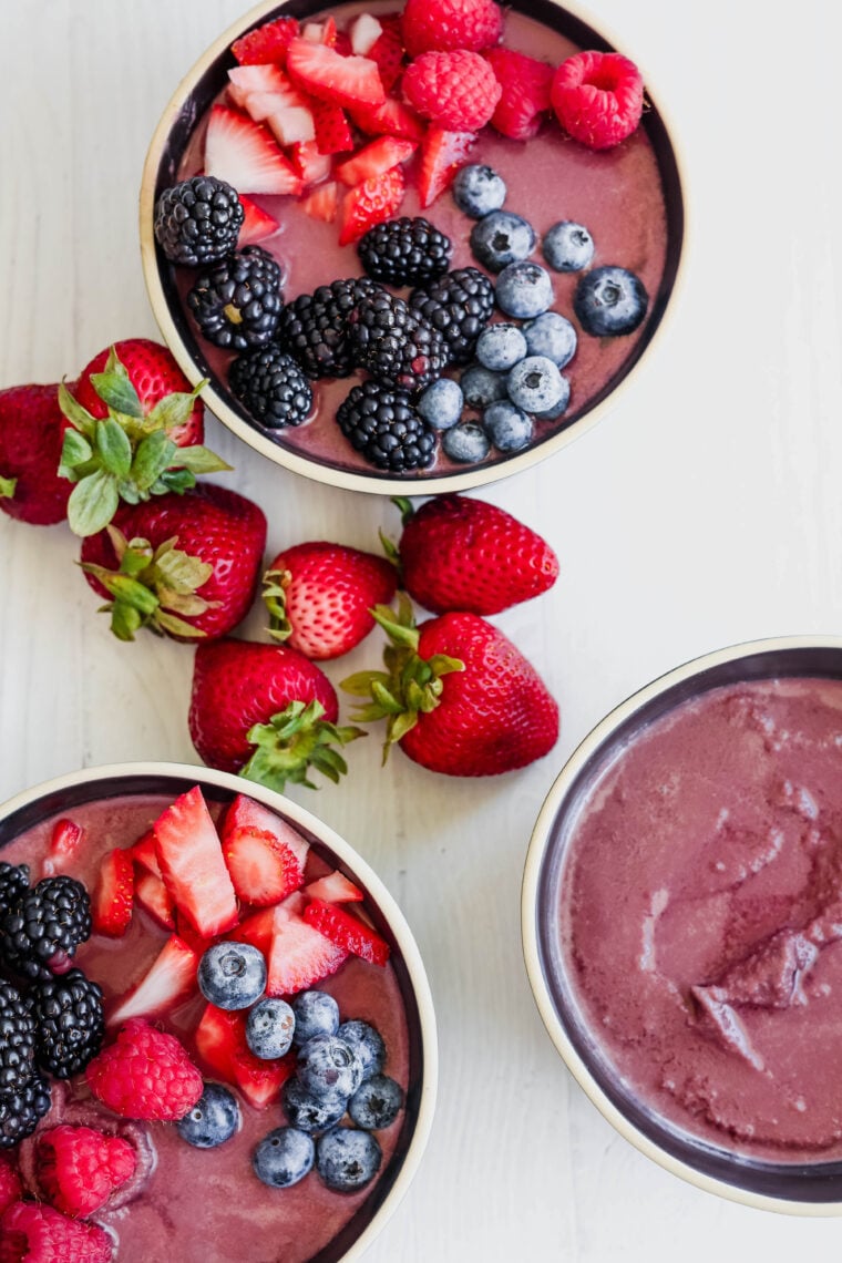 Three smoothie bowls—one’s a super colorful acai bowl—are loaded with fresh strawberries, blueberries, raspberries, and blackberries. They’re sitting on a white table next to some whole strawberries.