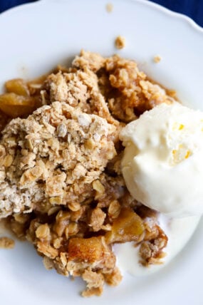A scoop of vanilla ice cream melts over a warm apple crumble on a white plate. The dessert’s got that sweet baked apple inside and a crispy oat topping—total comfort food!.