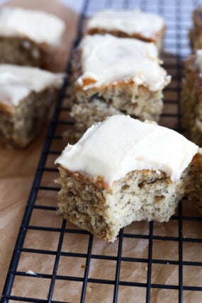 Here’s a close-up of some frosted banana bars on a black cooling rack, sitting on brown parchment paper. One of them’s got a big bite taken out—yum!.
