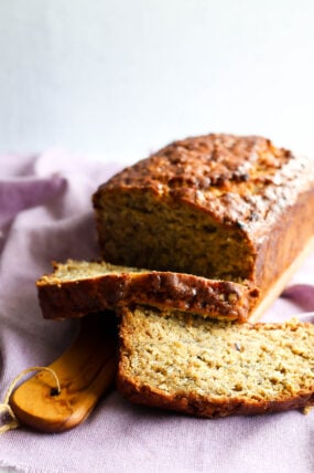 A loaf of banana bread with a perfect golden crust is chilling on a wooden board. Two slices are already cut, showing off that moist, yummy inside. Everything’s set on a cozy purple cloth.