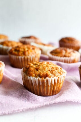 Some golden-brown muffins in white paper cups, hanging out on a light purple cloth with a soft blur in the background.