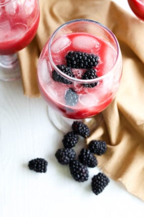 A glass of iced red blackberry drink topped with whole blackberries, sitting on a light surface with some fresh berries and a tan cloth nearby.