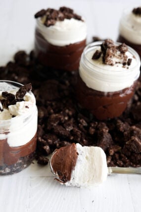 Four little jars packed with layers of chocolate pudding, whipped cream, and cookie crumbles are sitting on a cookie-covered surface. There’s also a spoon in front loaded up with pudding and cream, ready to dig in!.