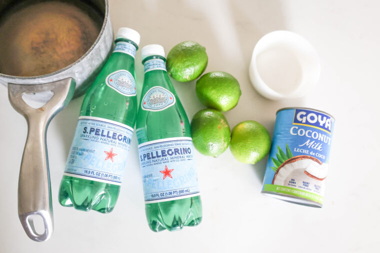 On a white surface, there&rsquo;s two bottles of San Pellegrino, four limes, a can of Goya coconut milk, a bowl of sugar, a saucepan, and a metal ladle all laid out.