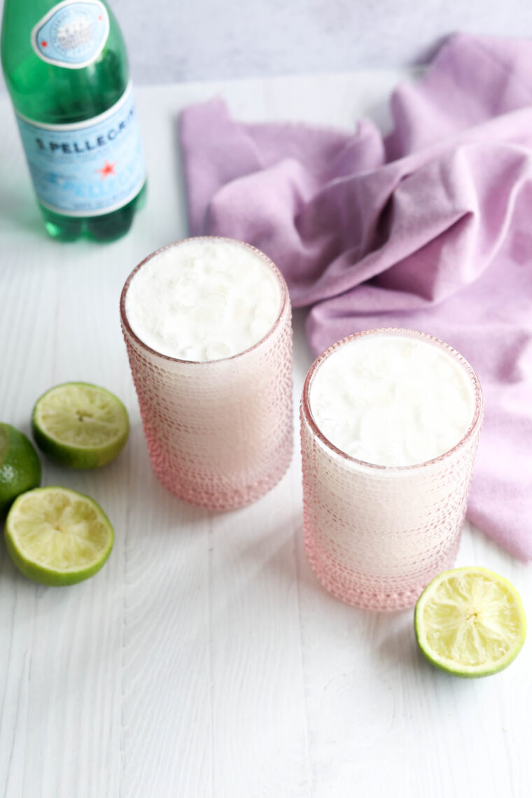 Two fizzy drinks in cool pink glasses sit on a white table, with lime halves, a purple cloth, and a bottle of sparkling water hanging out in the background.