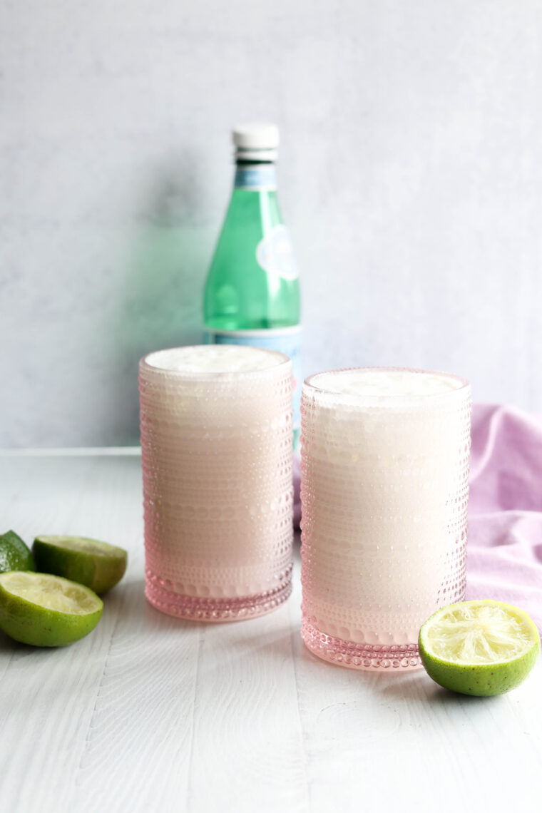 Two pink glasses with a frothy white drink are chilling on a white surface. Lime halves are next to them, a green bottle with a white cap is in the background, and there&rsquo;s a light purple cloth draped nearby.