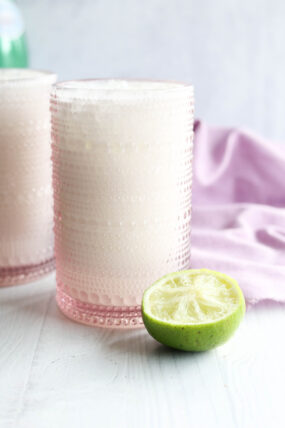 A pink, textured glass filled with a creamy white drink sits on a white surface next to a squeezed lime half, with a light purple cloth in the background.