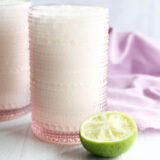 A pink, textured glass filled with a creamy white drink sits on a white surface next to a squeezed lime half, with a light purple cloth in the background.