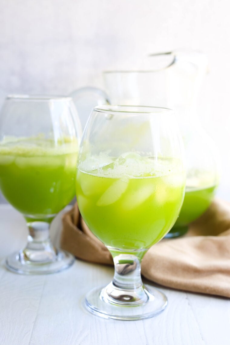 Two big glasses of iced green juice are sitting on a white table, with a glass pitcher and a tan cloth in the background.