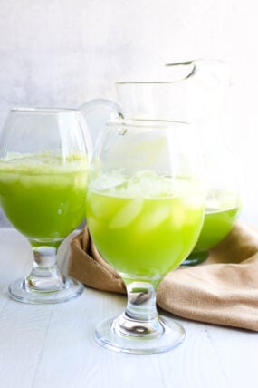 Two big glasses of bright green iced drinks sit in front of a pitcher with more, all on a white surface with a tan cloth underneath.
