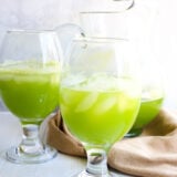 Two big glasses of bright green iced drinks sit in front of a pitcher with more, all on a white surface with a tan cloth underneath.