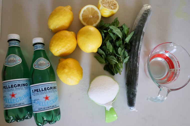 On a white surface: two bottles of San Pellegrino, four lemons plus one cut in half, some fresh mint, a whole cucumber, a cup of sugar, and a glass measuring cup filled with water.