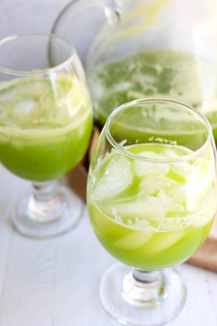 Two glasses with light green juice and ice are front and center, while a matching pitcher sits in the back. Everything’s set out on a pale surface—super refreshing vibes!.