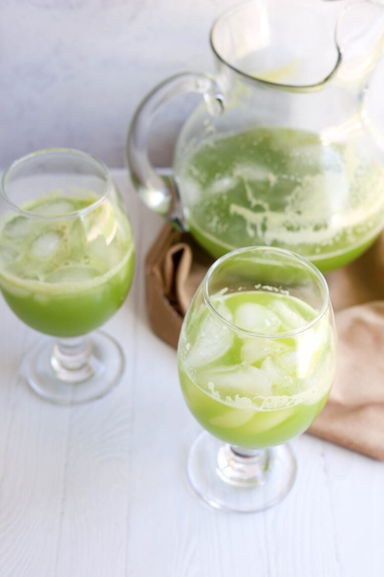 A glass pitcher and two goblets filled with icy, light green juice sit on a white surface next to a beige cloth. The drink looks super refreshing and frothy.