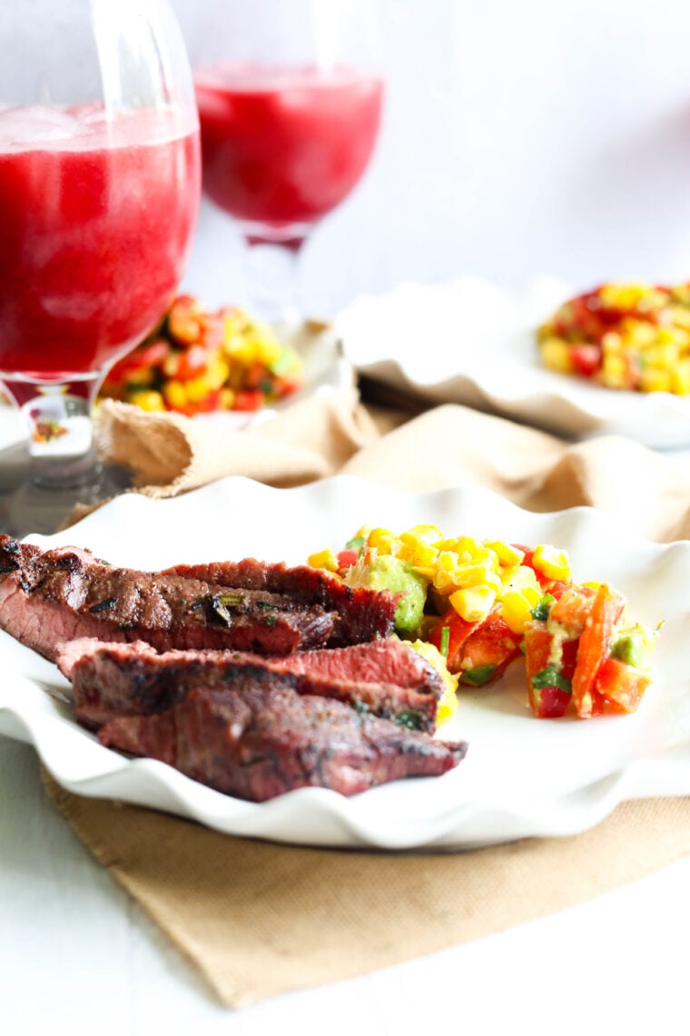 Three grilled steak slices and a vibrant veggie salad sit on a wavy white plate, with two glasses of red drink and some blurred plates in the background.