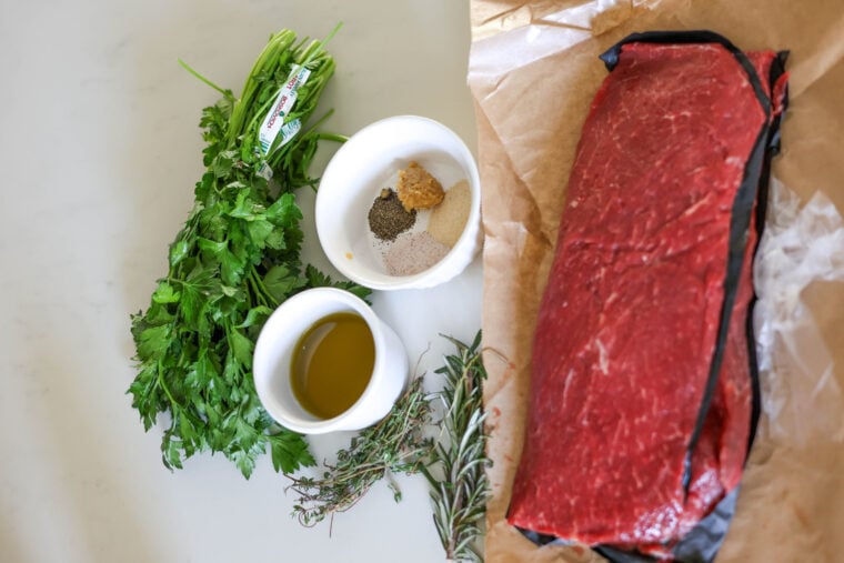 Fresh parsley, rosemary, and thyme, some olive oil, spices with mustard in a bowl, plus a big raw steak on brown parchment paper — all laid out on a white background.
