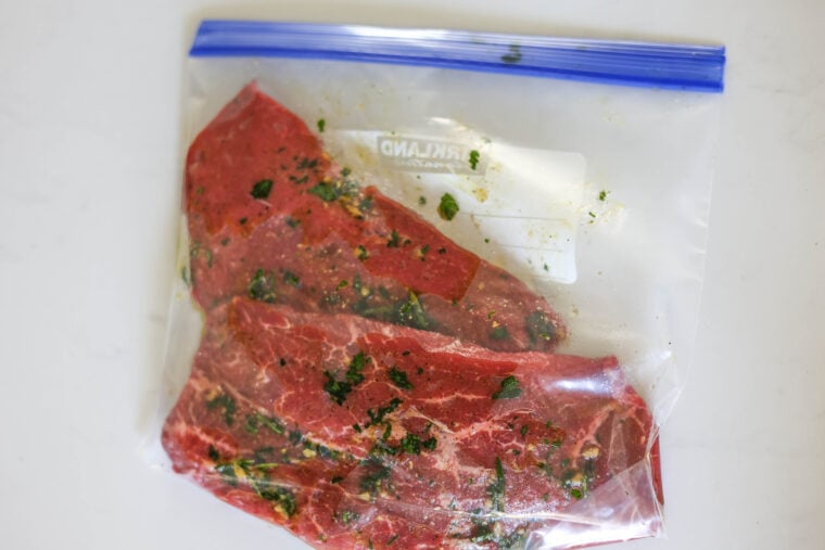 Two marinated beef steaks with herbs and spices, hanging out in a clear ziplock bag on a white table.