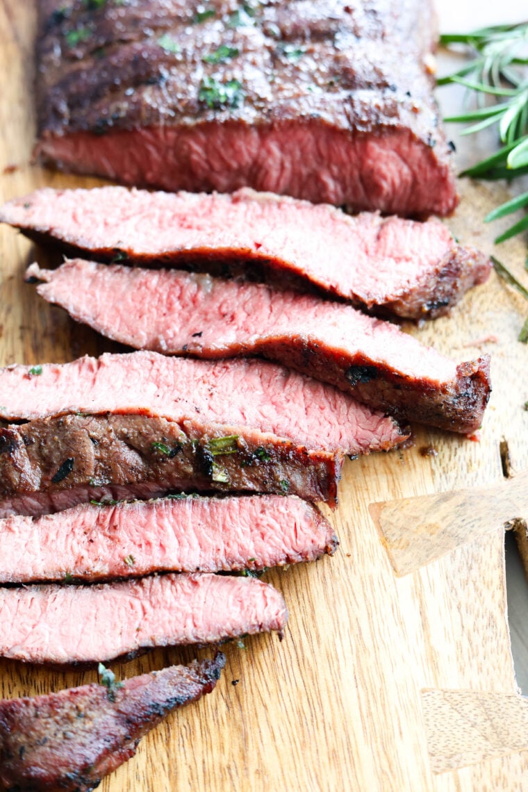 Grilled steak, perfectly pink and juicy in the middle, is sliced up on a wooden board with some fresh herbs on the side.